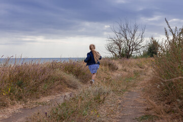 Naklejka na ściany i meble Adorable little girl in denim jacket back view on a hill with sea landscape view. Stylish child with long blonde hair on countryside cloudy sky background. Outdoor walking rural road trip faceless.