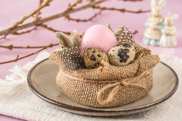 Handmade Easter breakfast decoration: jute fabric, pink Easter egg, natural quail eggs and feathers on a rustic plate, pinkish background