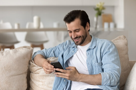 Smiling Millennial Caucasian Man Sit On Couch At Home Have Fun Using Modern Cellphone Gadget. Happy Young Male Relax On Sofa Look At Smartphone Screen Texting Or Messaging Online. Technology Concept.