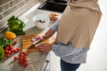 High angle view of a Muslim woman in hijab cutting tofu cheese and vegetables while preparing vegetarian salad.