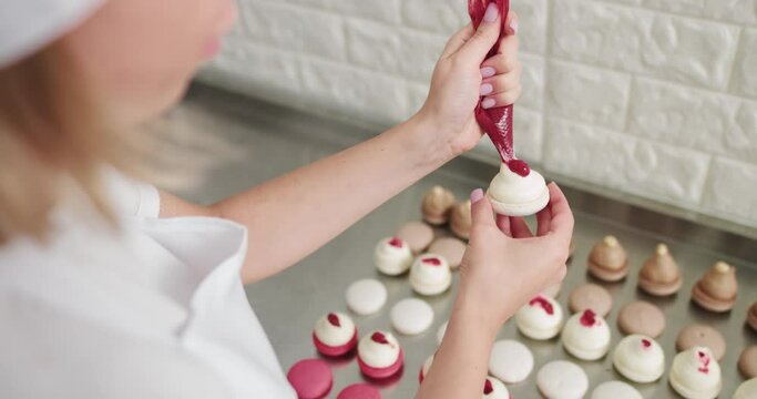 Shot of hands of female pastry chef holding white macaron with ganache and squeezing red fruit jam from bag. Beautiful pink macaroons fill with pink jam on a pink background. Nature pastry macaroons
