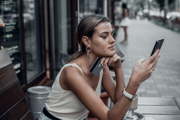 Young smiling woman shopping online using phone outdoors in cafe. A beautiful model looks at the phone and drinks coffee in the summer