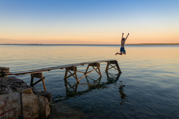 Boy Jumping from End of Dock into Calm Lake at Sunset at Family Cottage