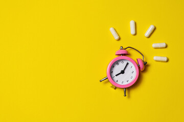 Pink alarm clock with pills in capsules on a yellow background.
