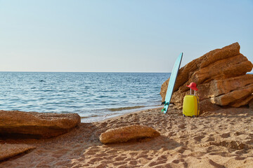 Yellow suitcase, surfboard and hat on sand. Sea view and rocky coastline. Summer time and holiday...