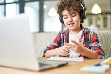 Joyful latin american little boy wearing headphones, looking at the screen of a laptop while...