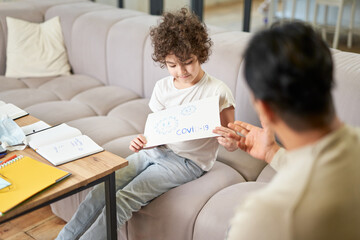 Fototapeta premium Stay home together. Happy latin boy showing the drawing Covid 19 to his dad while spending time with his father, playing in the living room at home