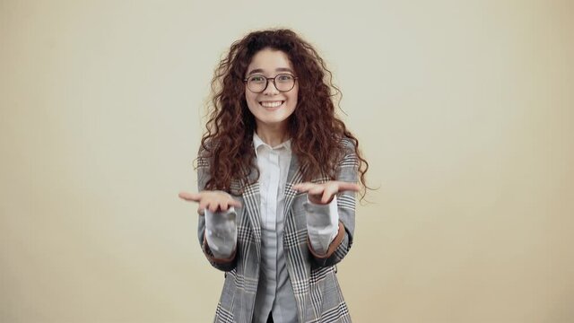 The amazed, cheerful and enthusiastic young woman, with her curly hair in a gray jacket and white shirt, with glasses posing isolated on a beige background in the studio. The concept of people's