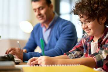Obraz premium Happy teenaged latin boy in glasses making notes while sitting at the desk together with his father and doing homework indoors
