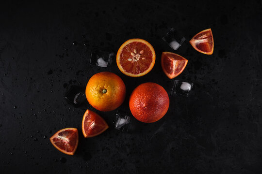 ripe red oranges. whole, half and triangular slices lie scattered diagonally on a dark background with ice cubes and water droplets. top view with side light. dark artistic image for creative mockup