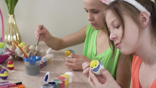 Two girls decorate Easter eggs together and talk. Eggs, paints, and markers are on the table. Easter holiday.
