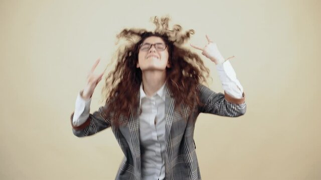 The crazy young woman dances rock with curly hair in a gray jacket and white shirt, with glasses posing isolated on a beige background in the studio. The concept of people's lifestyle. With his