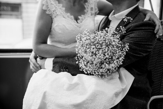 Black and white image of bride sitting on lap of groom in a bus. Bride holding Gypsophila bouquet