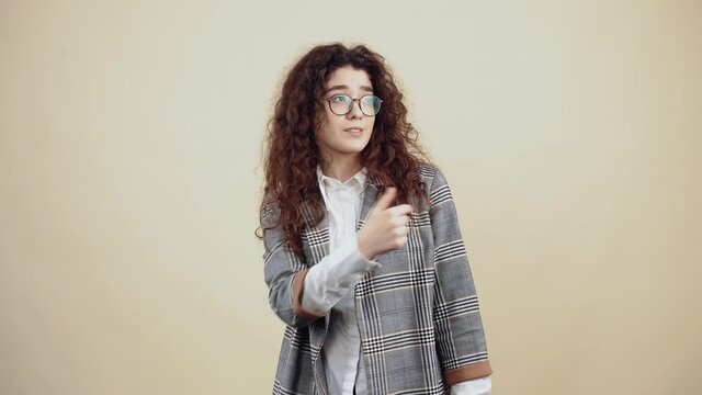 Hey you, look there. Beautiful young woman with curly hair in gray jacket and white shirt, with glasses posing isolated on beige background in the studio. The concept of people's lifestyle. Point your