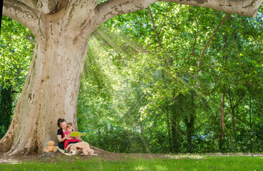 Mother reading a book with her cute little child under an old plane tree in the park. Funny and educative outdoor activity for children, in sunny summer day