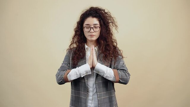 The calm young woman, balanced with her hands together, prays and meditates with her eyes closed Cretaceous in gray jacket and white shirt, with glasses posing isolated on a beige background in the