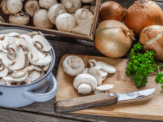 Raw mushrooms, whole and sliced, lined from a wooden board box and placed in a metal pot. Light onions and parsley leaves.