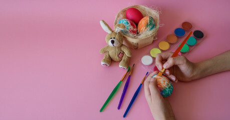 Child`s hands paint Easter egg next to the fluffy toy rabbit, the nest with handmade colored eggs, multicolored paints and art brushes on a pink background. 