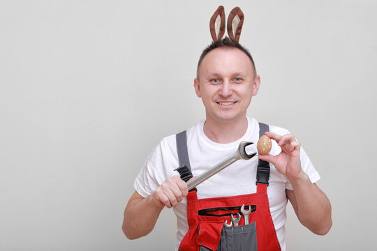 Easter holiday, celebration concept. funny smiling engineer worker or mechanic is wearing rabbit ears on head, holding wrenches and painted egg white background. celebrating orthodox day