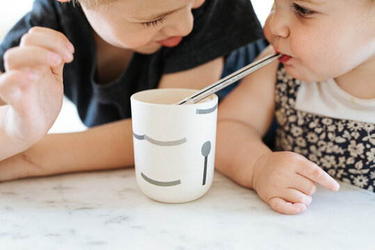 brother helping sister drink from cup with plastic straw