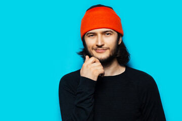 Studio portrait of confident young man wearing orange hat and black sweater on blue background.