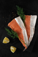 sliced fresh raw salmon lying on a stone cutting board with lemon wedges, dill and spices on a black textured concrete background. top view. beautiful sea food concept
