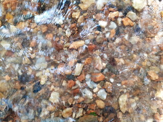 Ripples in the river flow through many stones, colorful summer to bright sunlight. The background shiny pebble, with A bottom of stream In an abstract, form Of pebbles below water surface.