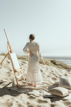 Young woman painting on an easel at the coast in the sand