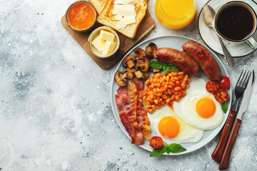Full English breakfast on a plate with fried eggs, sausages, bacon, beans, toasts and coffee on light stone background. With copy space. Top view