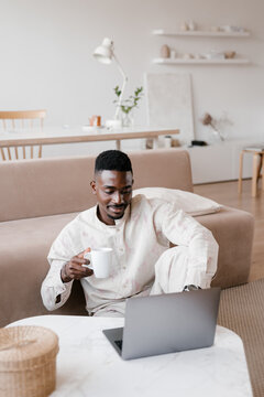 Cheerful ethnic man with cup of drink using laptop at home