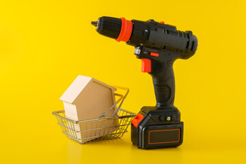 Modern black cordless screwdriver, drill with rechargeable battery and wooden house in shopping basket on yellow background, copy space for text, construction or renovation concept