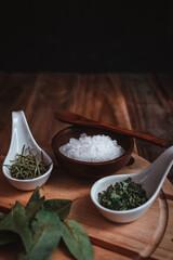 Ceramic posillo with salt, accompanied by two white posillos with oregano and rosemary, bay leaves, on a wooden board, dark and rustic background