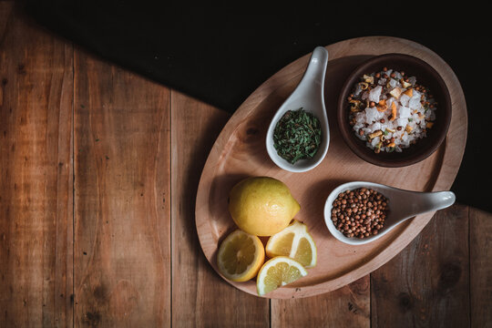 Ceramic posillo with salt and spices, lemon and coriander seeds, accompanied by spices, oregano and cut lemon, on wooden board and dark rustic background