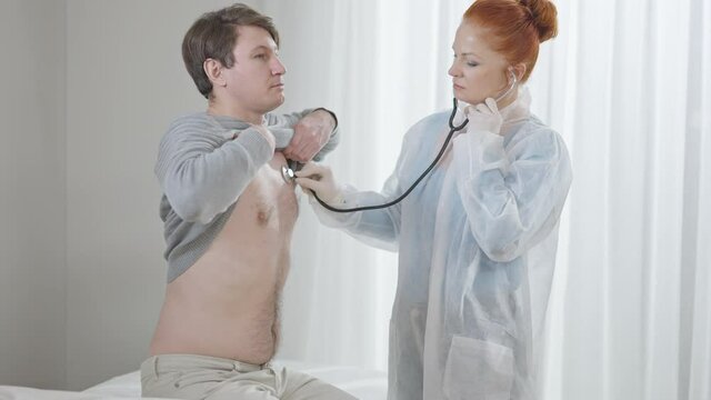 Concentrated woman listening internal sounds as man sitting on medical couch in hospital ward. Caucasian doctor using stethoscope for chest auscultation as patient inhaling and exhaling.