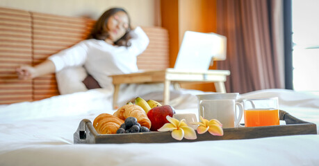 Obraz na płótnie Canvas Breakfast in a tray on the bed in the luxury hotel room in front of an Asian businesswoman traveler work with a laptop, healthy food concept