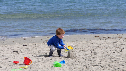 Red headed boy playing with Bucket spade and Digger on a sandy beach 