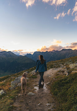 A young woman in a blue jacket walking with her dog in the mountains during the sunset.