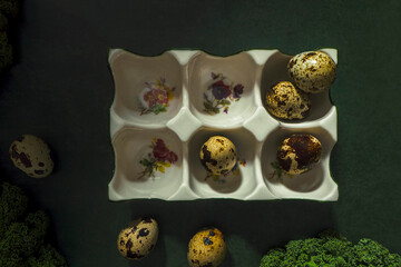 Small spotted quail eggs in the white porcelain floral egg tray on a dark green background. Creative still life and raw photography. Elegant art of healthy food, flat lay photography.