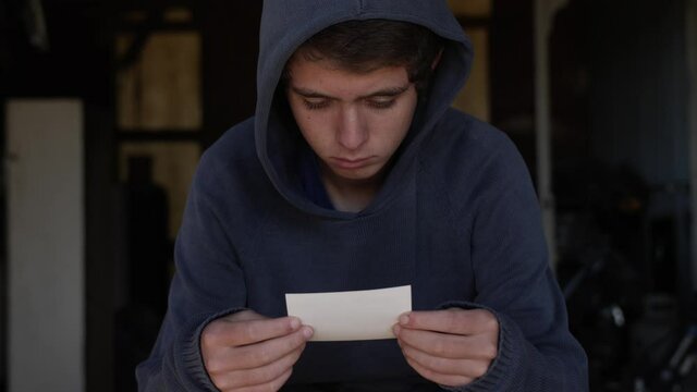 A sad and depressed looking teenage boy wearing a blue hooded shirt sits alone in a dirty garage looking at a picture. Filmed in 60 fps.
