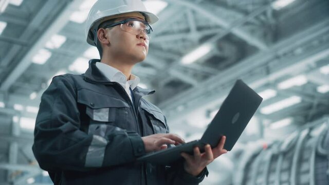 Portrait of a Professional Heavy Industry Asian Engineer/Worker Wearing Safety Uniform and Hard Hat Uses Laptop Computer. Confident Chinese Industrial Specialist Standing in a Factory Facility.