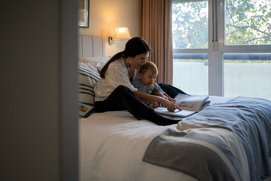 Young Mother Reading A Book To Her Baby On The Bed.
