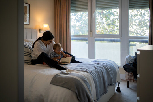 Mom Reading A Book To Her Baby On The Bed.