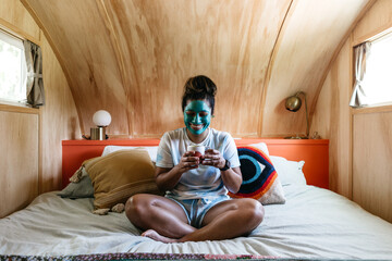 A woman sitting in bed with a cup of coffee and a cleansing face mask on.