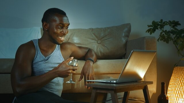 Young African American man using laptop, drinking red wine from glass, looking at the screen, talks during a video conference