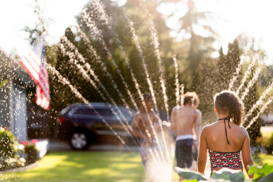 Girl in swimsuit watches brother play in sprinkler