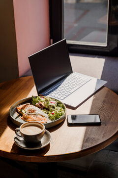 Laptop phone coffe and breakfast dish on top of wooden table in a cafeteria