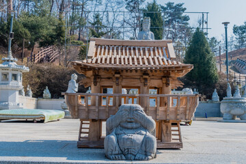 Sitting Buddha with hands over eyes in front of miniature wooden pavilion in plaza