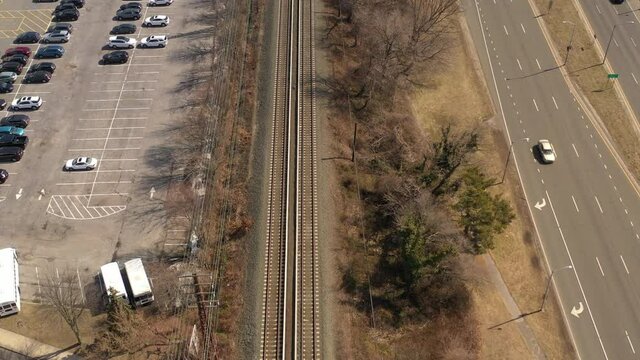 A top down shot of empty train tracks on a sunny day. On the left is a parking lot and on the right is trees, grass and a highway with light traffic. The camera, tilted down dolly in over the tracks.