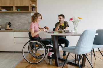 Young positive disabled woman in a wheelchair having a lunch with her boyfriend at home