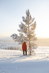 A man in a red jacket stands under a tree in the sun